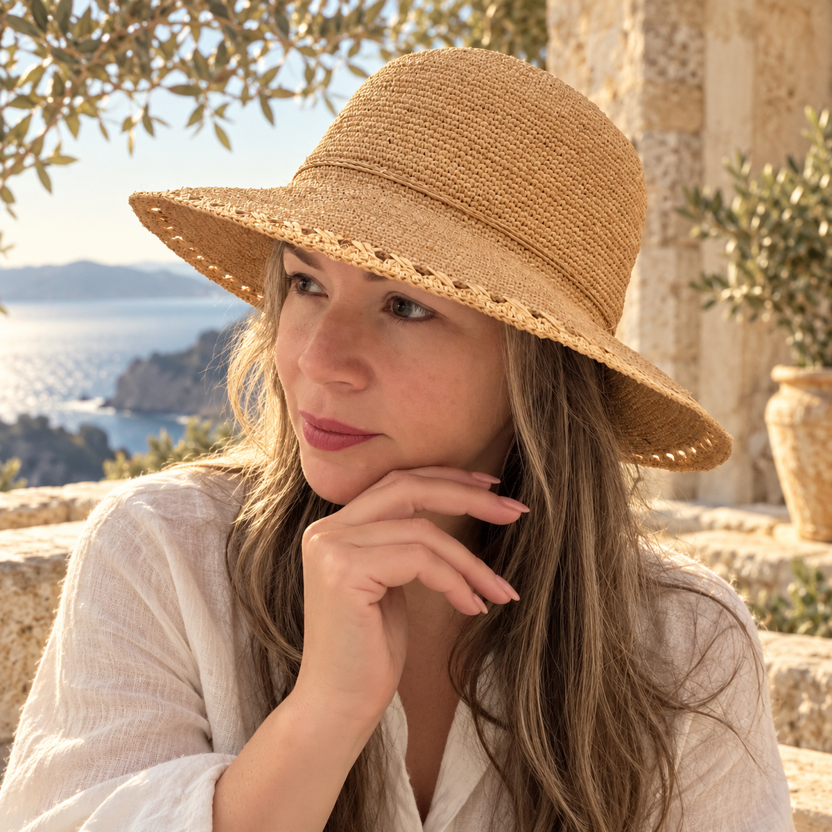 Woman wearing Alba natural raffia wide-brim bucket hat on a Mediterranean stone terrace by the sea.
