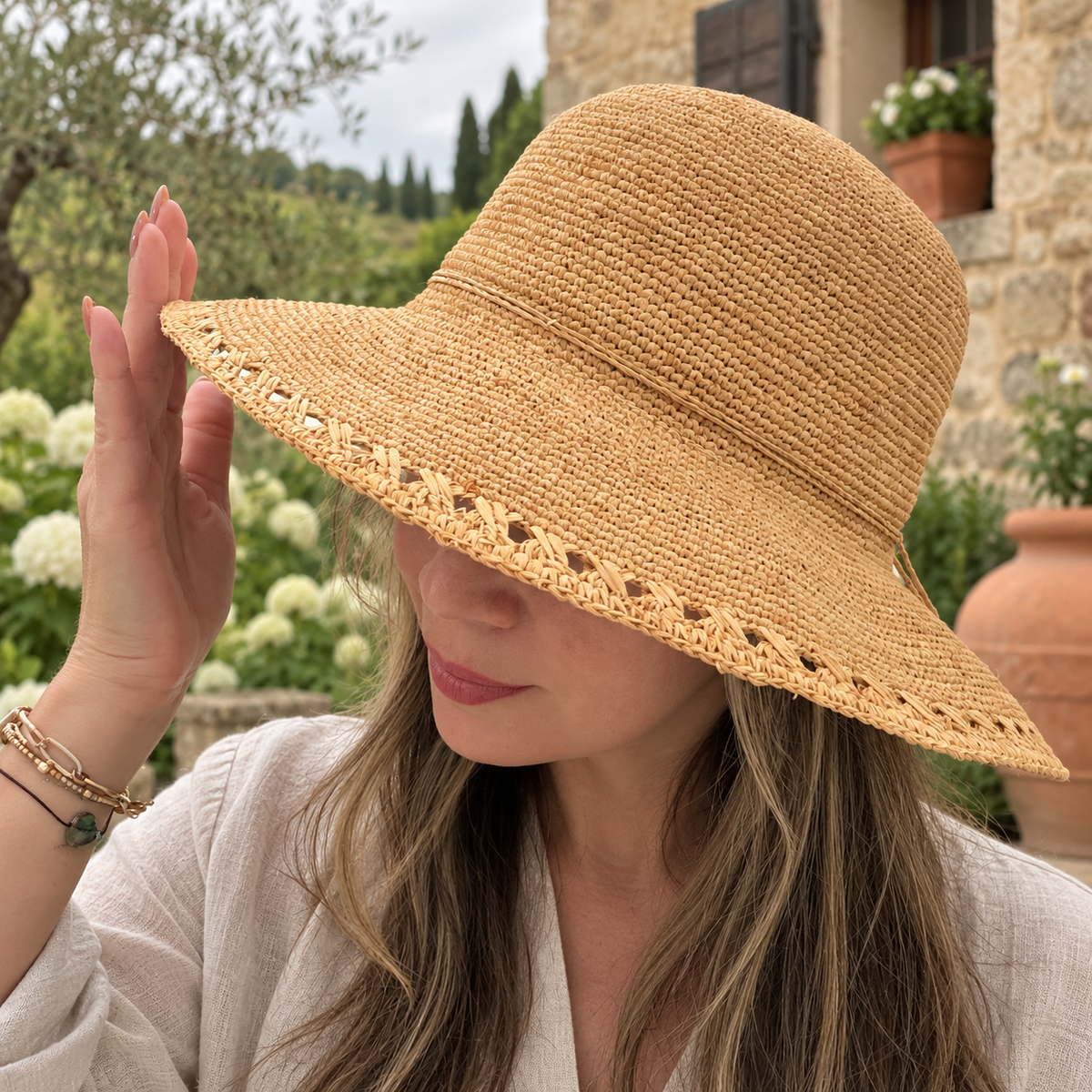 Woman wearing Alba natural raffia wide-brim bucket hat in a rustic villa garden.