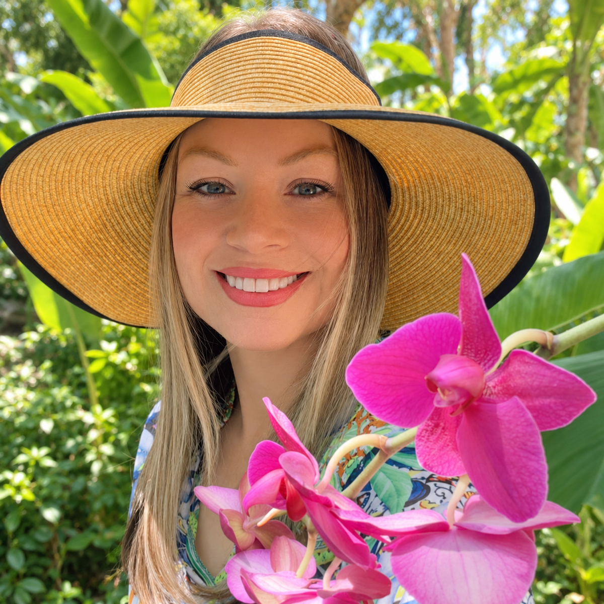 Woman wearing the Caro sun visor in a tropical garden beside pink orchids.