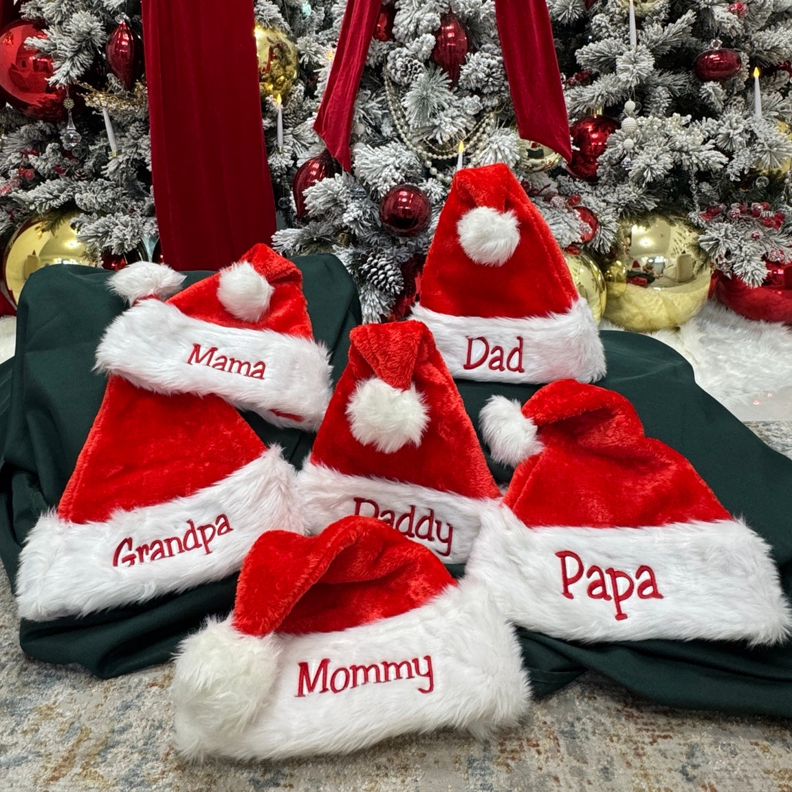 Six Santa hats with family names embroidered in red and Christmas trees behind them