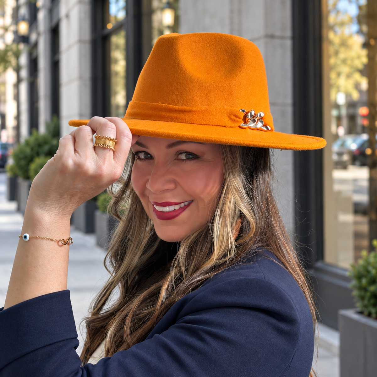 Woman wearing the orange Aris fedora with a metallic chain accent and a navy blazer on a city street.