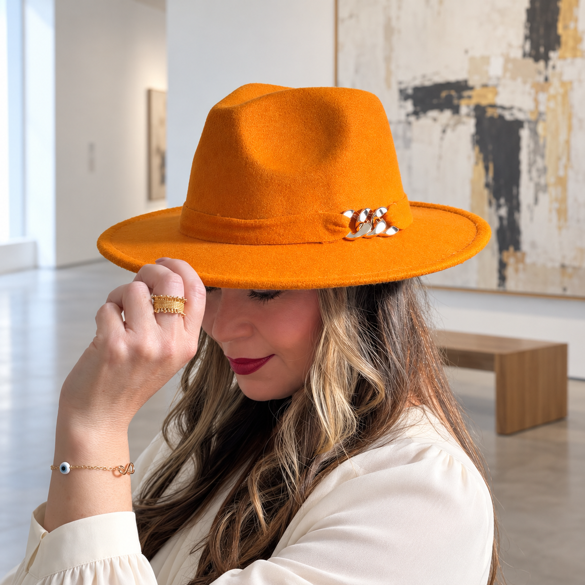 Woman wearing the orange Aris fedora with a metallic chain accent and an ivory blouse in an art gallery.