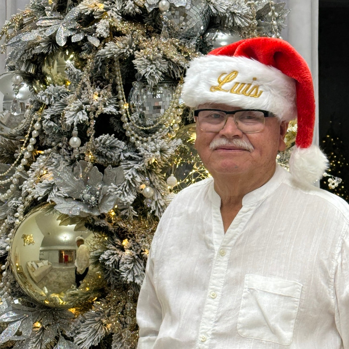 Luis gold embroidered Santa hat beside a Christmas tree