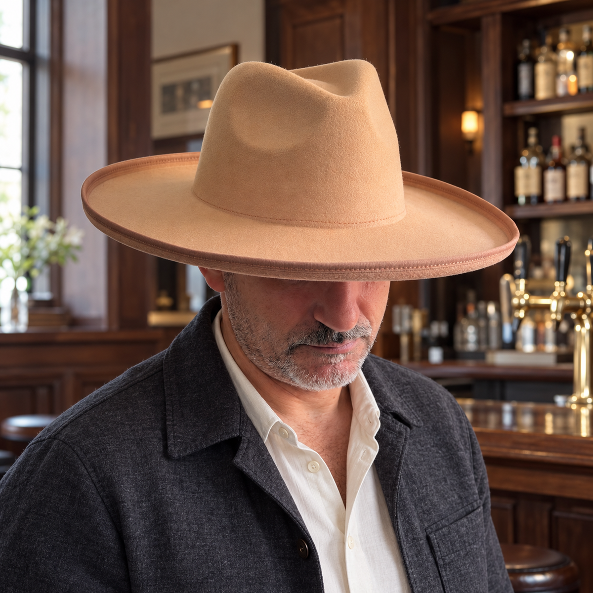 Man wearing the camel Orfeo vegan felt pencil roll brim fedora with a charcoal jacket in a wood bar.