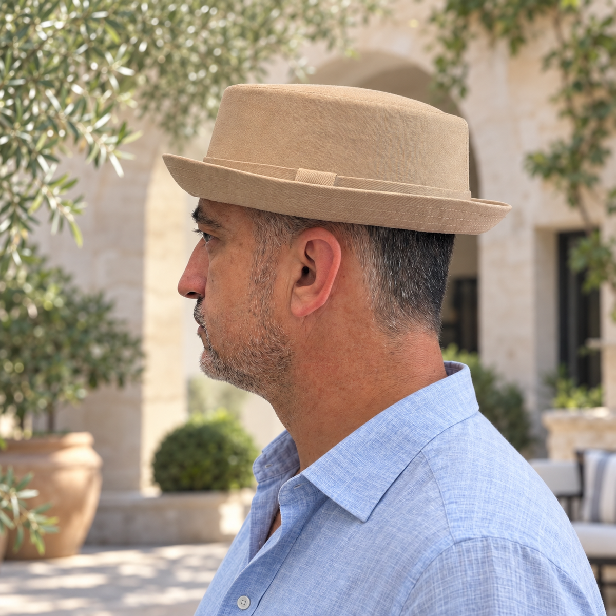 Man wearing the Oxford khaki cotton pork pie hat with a blue linen shirt in a Mediterranean courtyard.