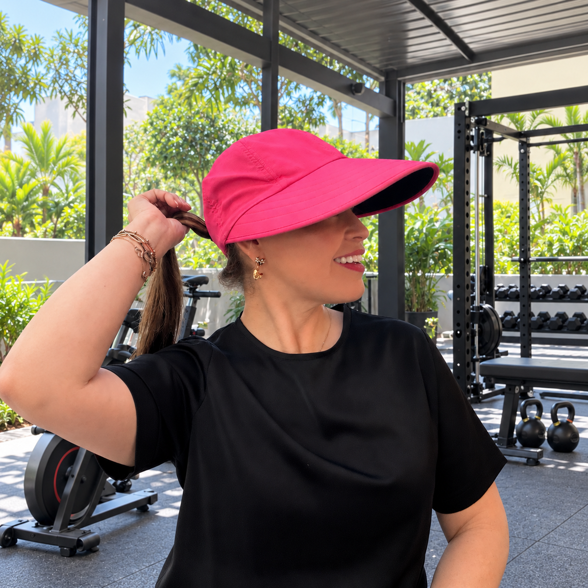 Woman wearing the hot pink Pia ponytail visor hat in an open-air gym, showing its wide brim and ponytail opening.