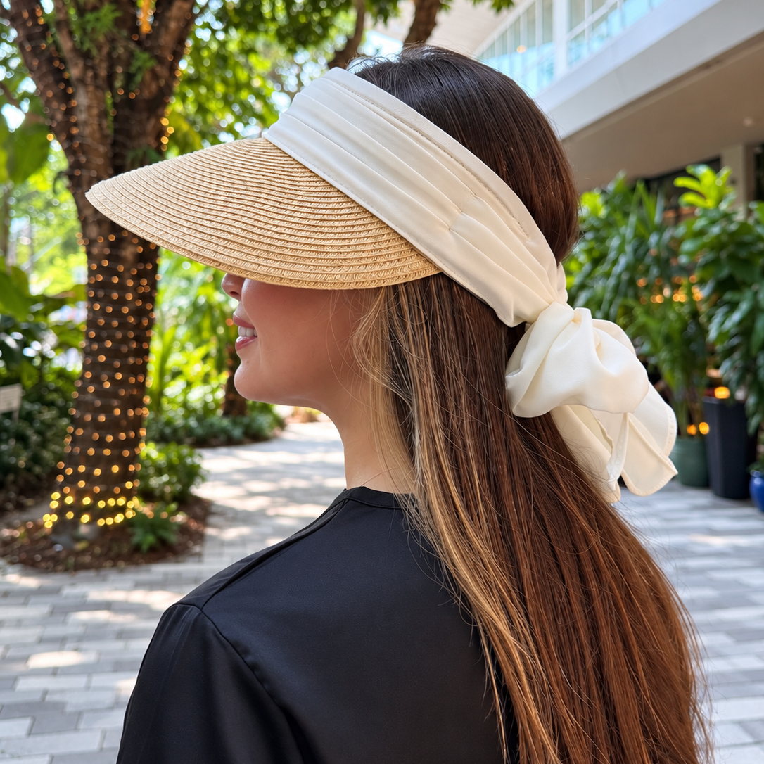 Woman wearing Vero natural sun visor with an ivory chiffon bow, seen in profile on a tropical promenade.